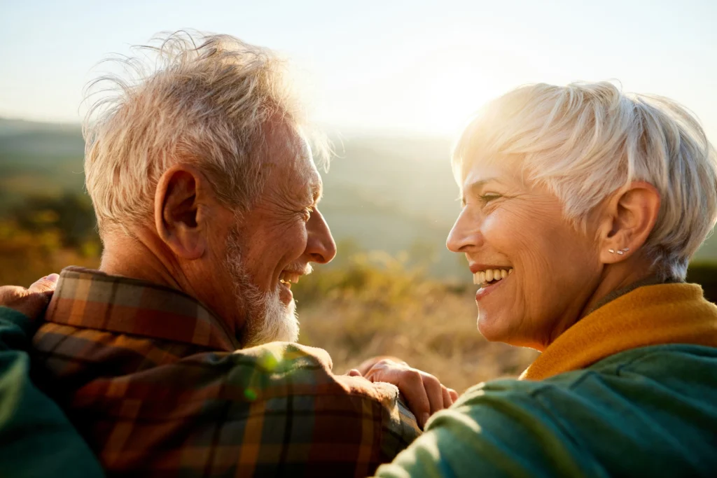 early-70s, independent living couple looks at each other, smiles, while seated on a hill in Phoenix, AZ.