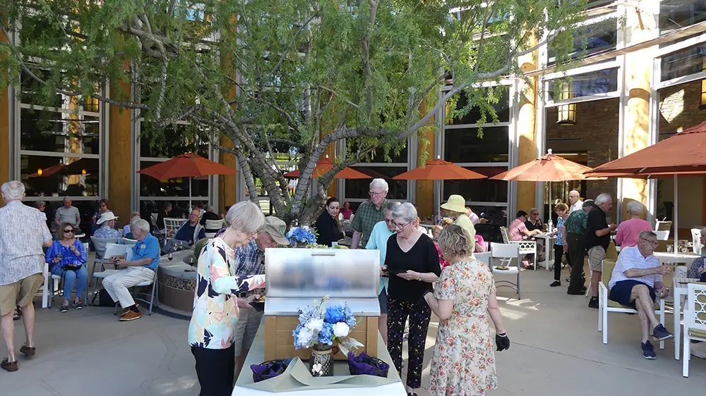 A lively group of Sagewood residents gather at an outdoor patio buffet and dining area under orange umbrellas, enjoying the vibrant social opportunities offered at luxury retirement communities.